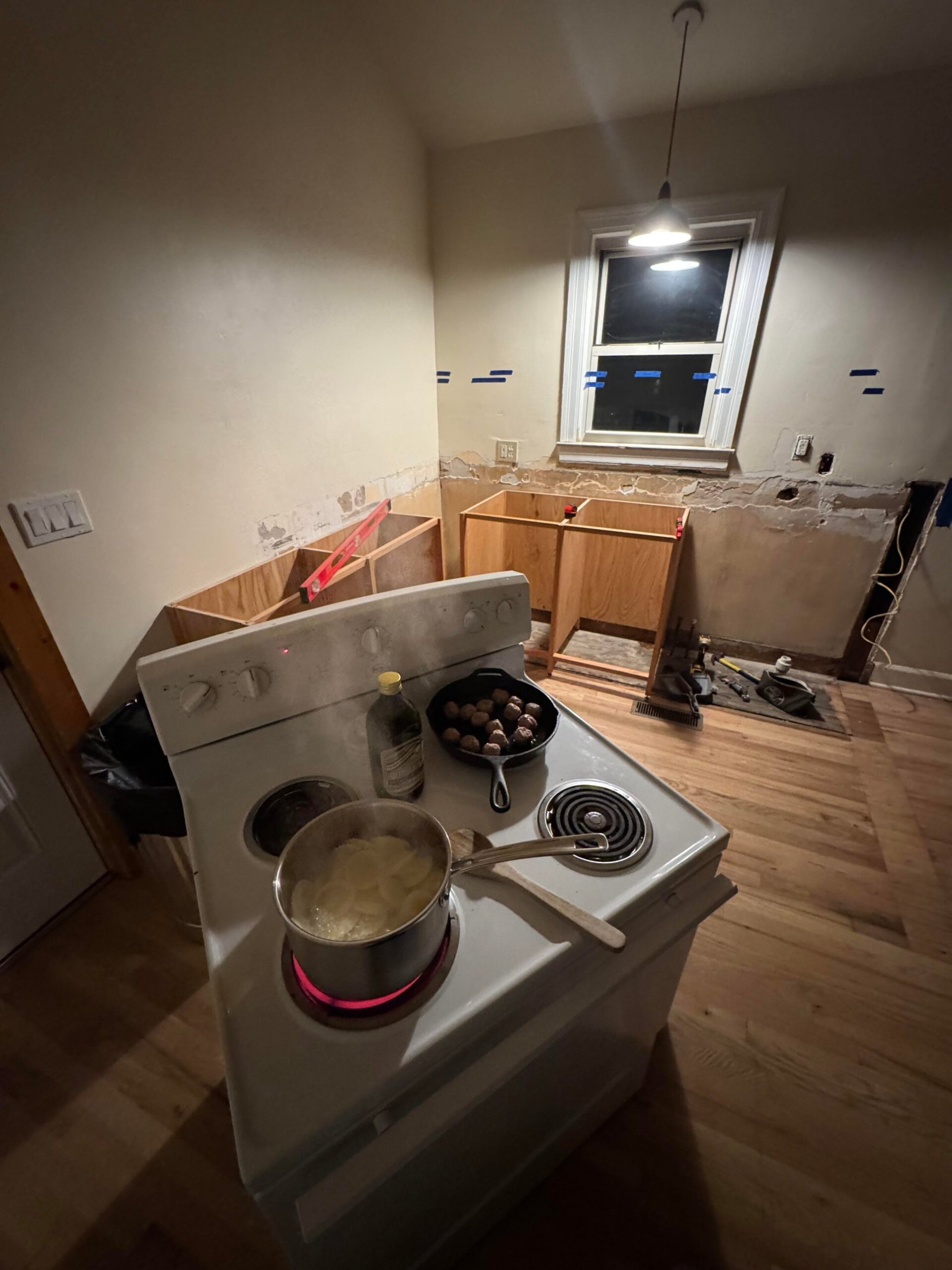 Overhead view of a white electric range with a cast iron pan of meatballs and a pot of boiling potatoes, with raw plywood cabinet frames being built in the background.