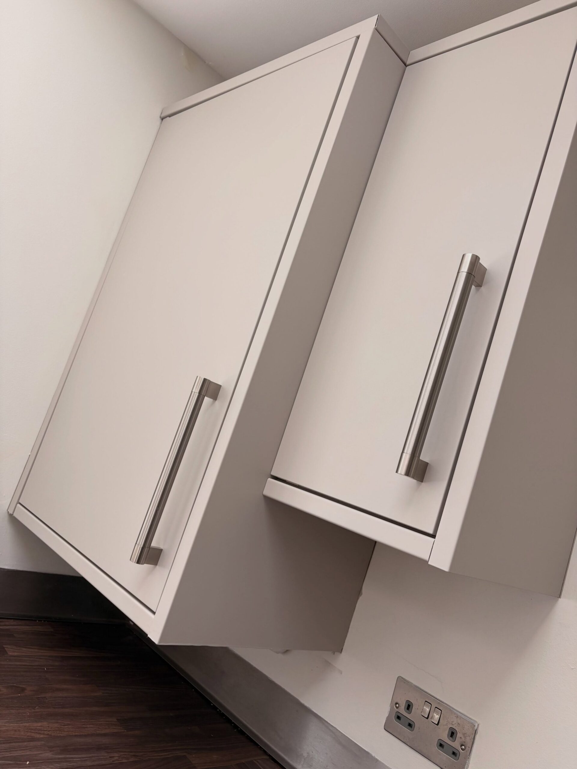 Close angled view of two sprayed Purbeck Stone cabinet doors meeting at a corner, each with a shorter brushed nickel bar handle, matte finish catching the ceiling light.