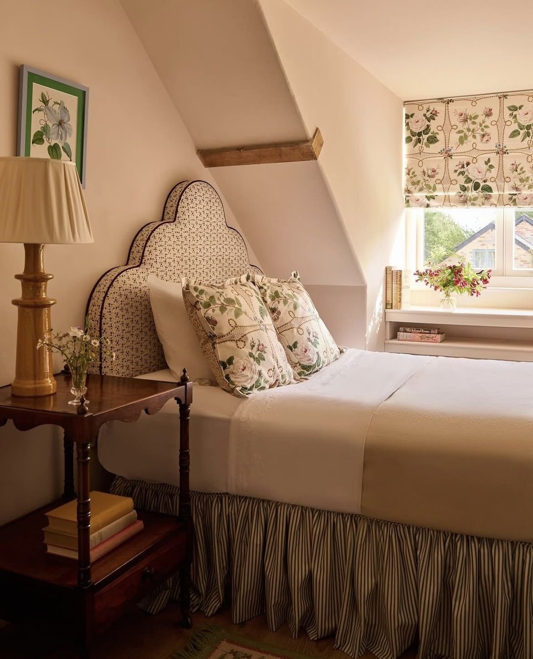 Cottage style attic bedroom with scalloped polka dot headboard, floral rose pillows, ticking stripe bedskirt, antique nightstand with brass lamp, and floral Roman shade