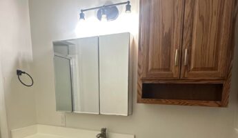 Small bathroom before the remodel, with orange oak cabinets, a white cultured marble vanity top, chrome fixtures, a white toilet to the right, and a tall oak storage cabinet mounted above it.