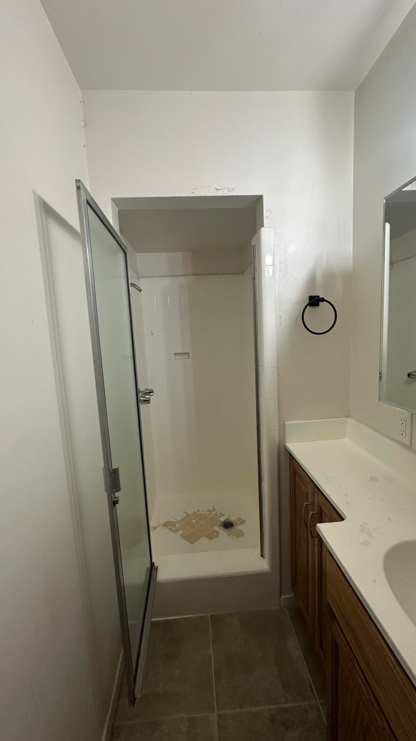 A tiny white tile shower stall with the frosted door swung open, missing tiles visible on the floor near the drain, and the vanity in the corner of the frame.