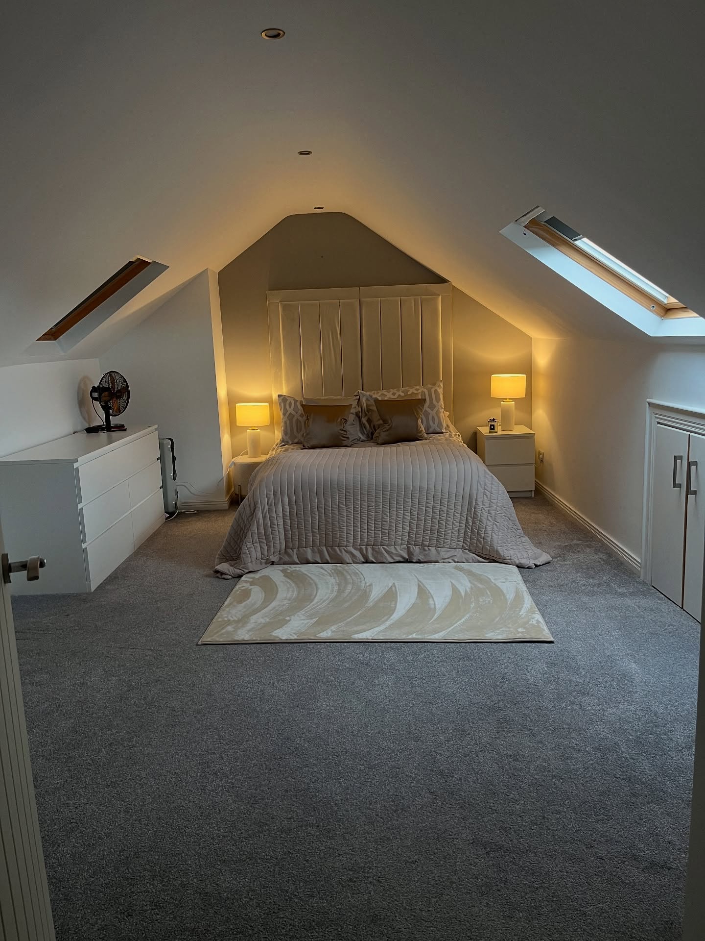 Clean cream attic bedroom with gray carpet, two angled skylights, upholstered paneled headboard reaching toward the peak, matching white bedside tables with lamps, and a dresser along the knee wall