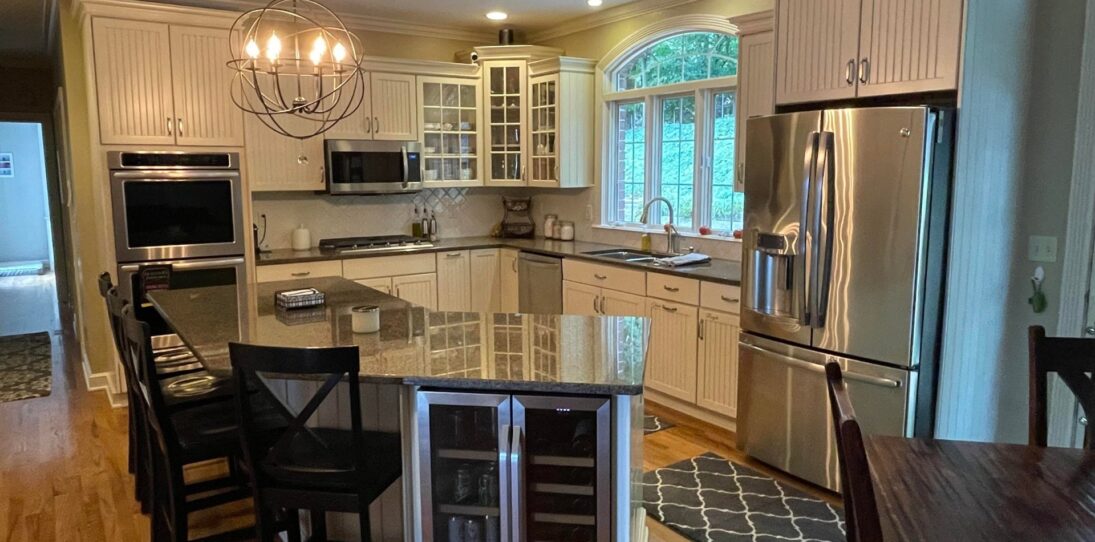 Cream beadboard kitchen with granite counters, an angled island with a built-in wine fridge, a stainless French-door refrigerator, double wall ovens, and an orb chandelier over the eating area.