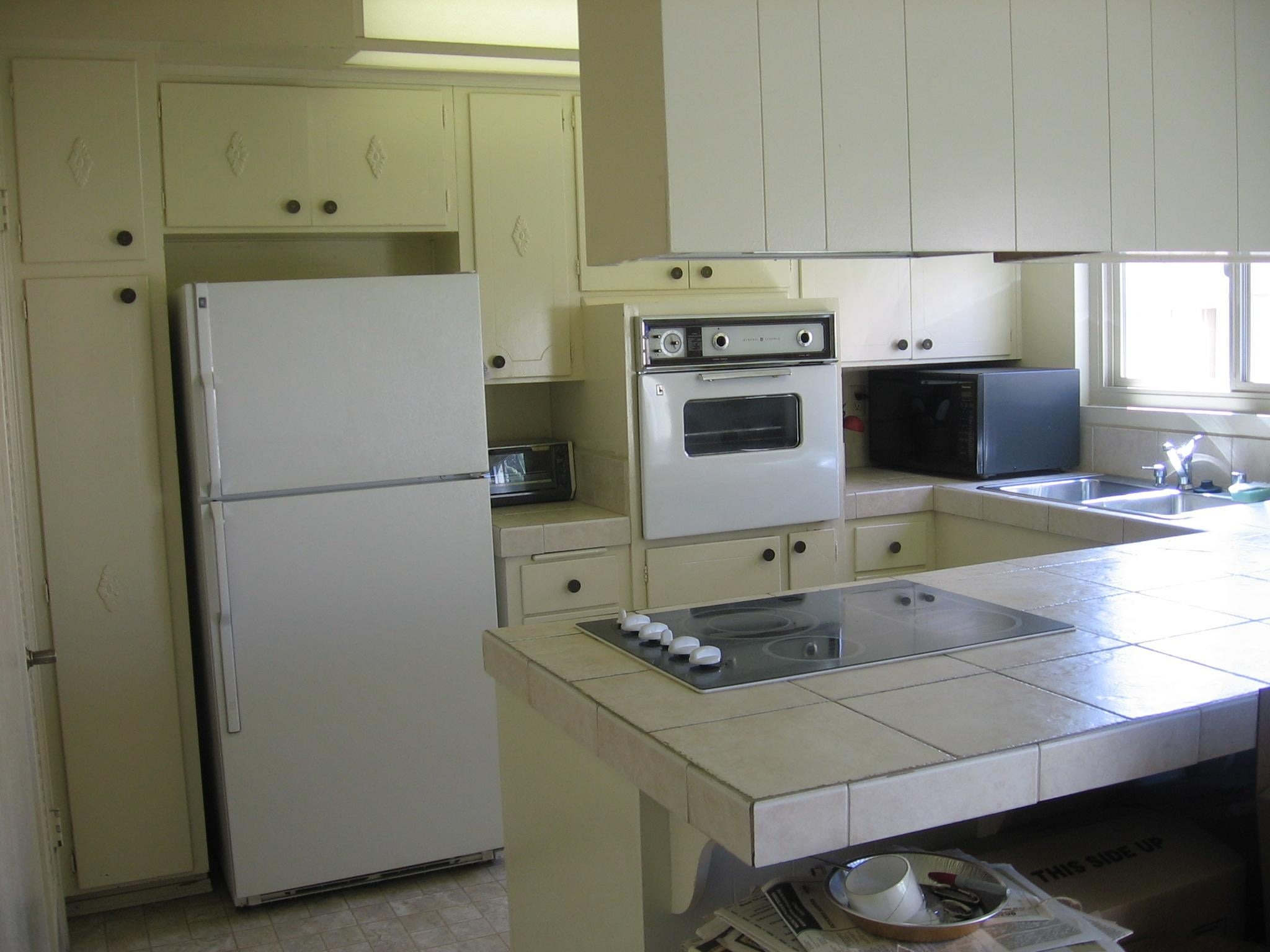 U-shaped 1966 kitchen with cream painted cabinets, a vintage white GE wall oven in the middle of the run, a smooth cooktop set into a tiled peninsula, and tile counters.