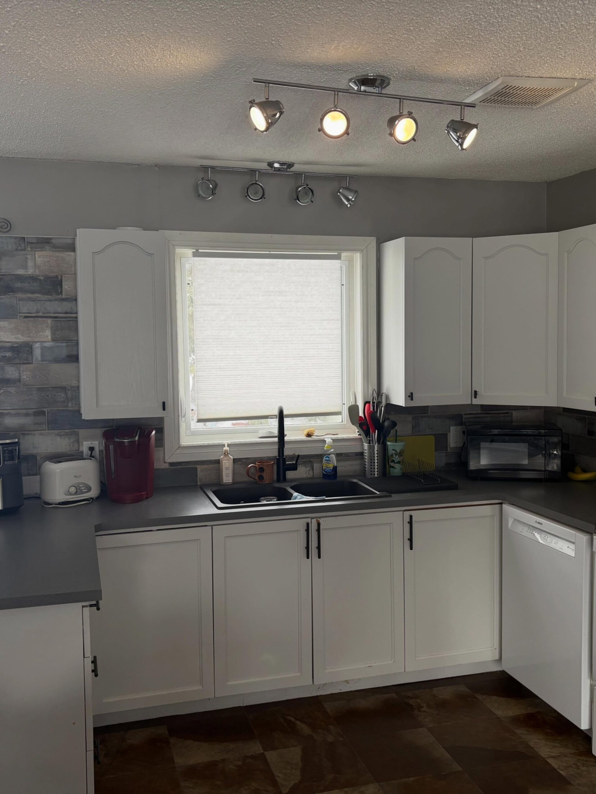 Dated kitchen with cream arched cathedral upper cabinets, chrome track lighting on a popcorn ceiling, a dark gray laminate counter, a black double sink, and a rustic multi-tone subway tile backsplash on the sink wall.