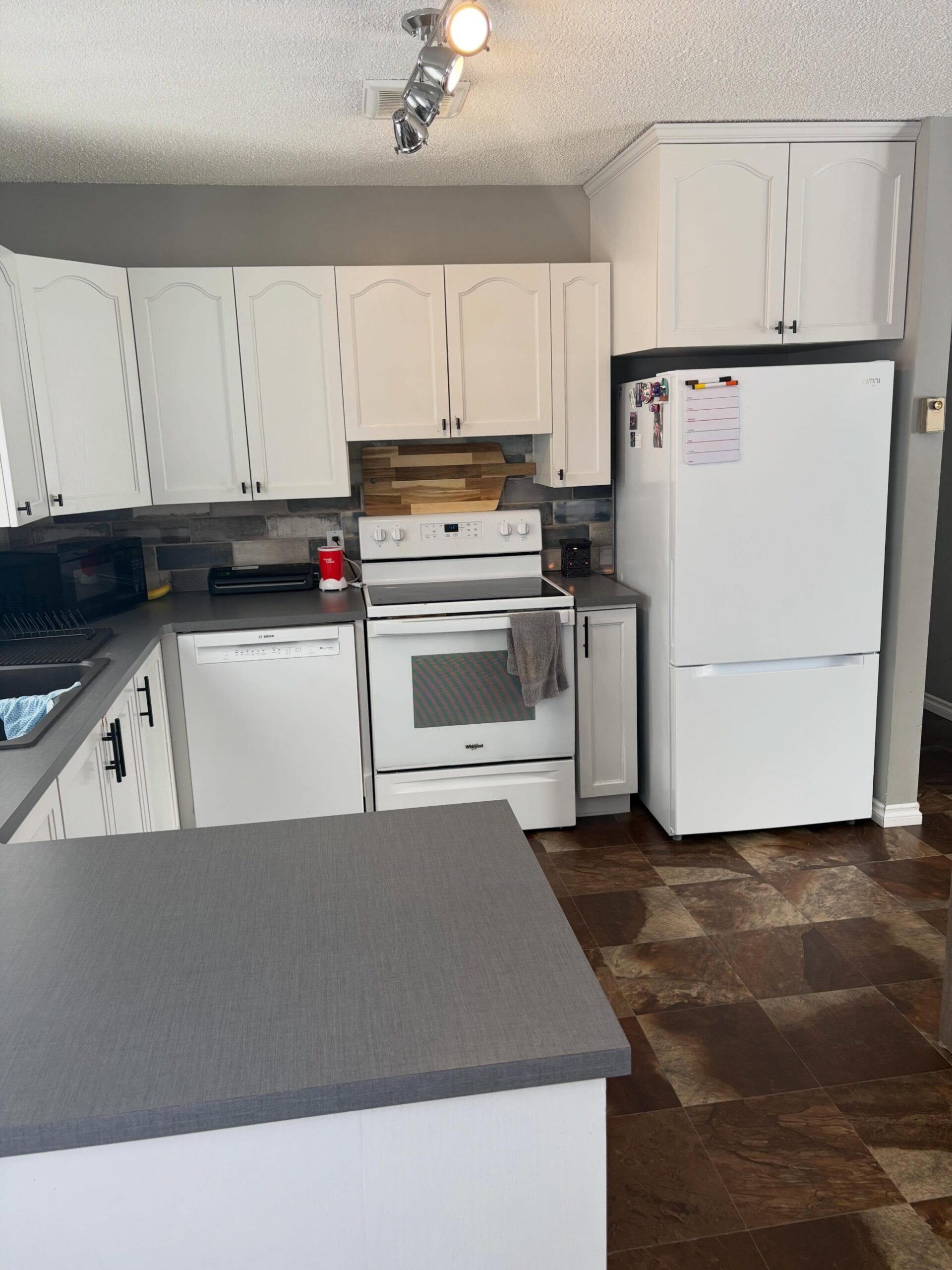 Range wall before refacing showing cream arched cathedral upper cabinets with black knobs, a white Whirlpool stove, a white fridge, dark brown tile flooring, and the same rustic blue-and-brown subway tile backsplash running behind the range.