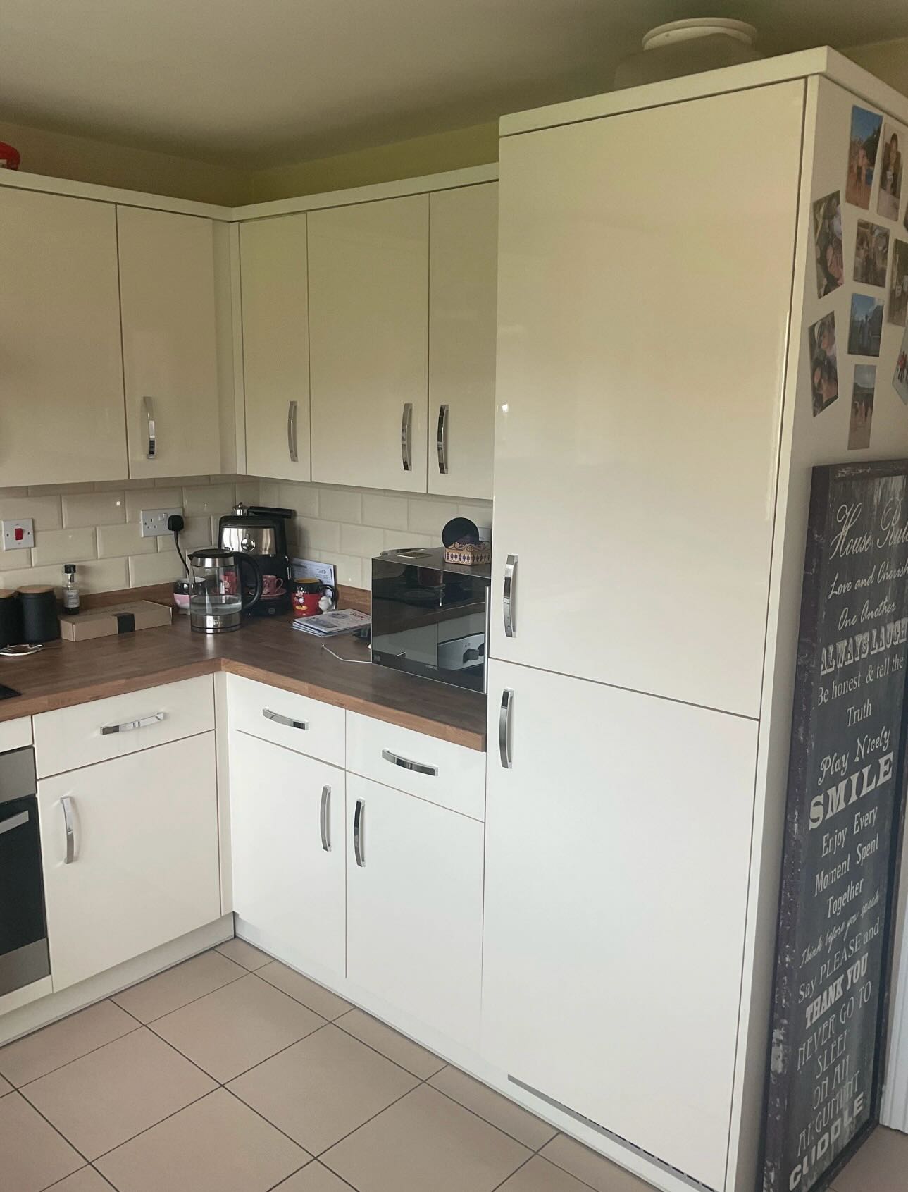 Cream high-gloss kitchen pantry corner with tall pantry cabinet, chrome bar handles, white subway tile backsplash, dark walnut-effect counter, and a wall of family photos beside a vintage house rules sign.