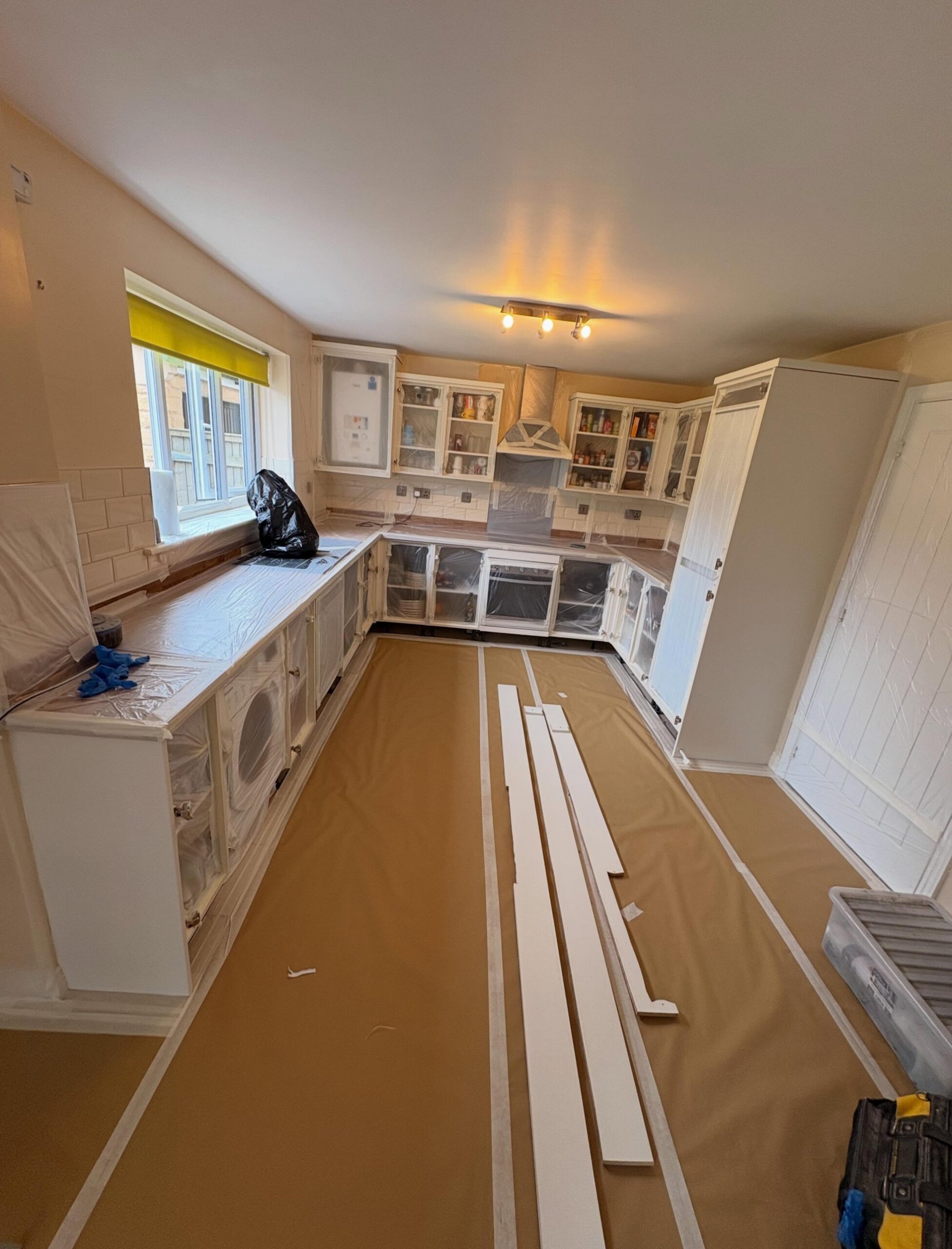 Cream kitchen in the middle of a respray with counters wrapped in plastic sheeting, floor covered in brown paper, cabinet doors taped off, and loose doors laid out on the floor ready for spraying.