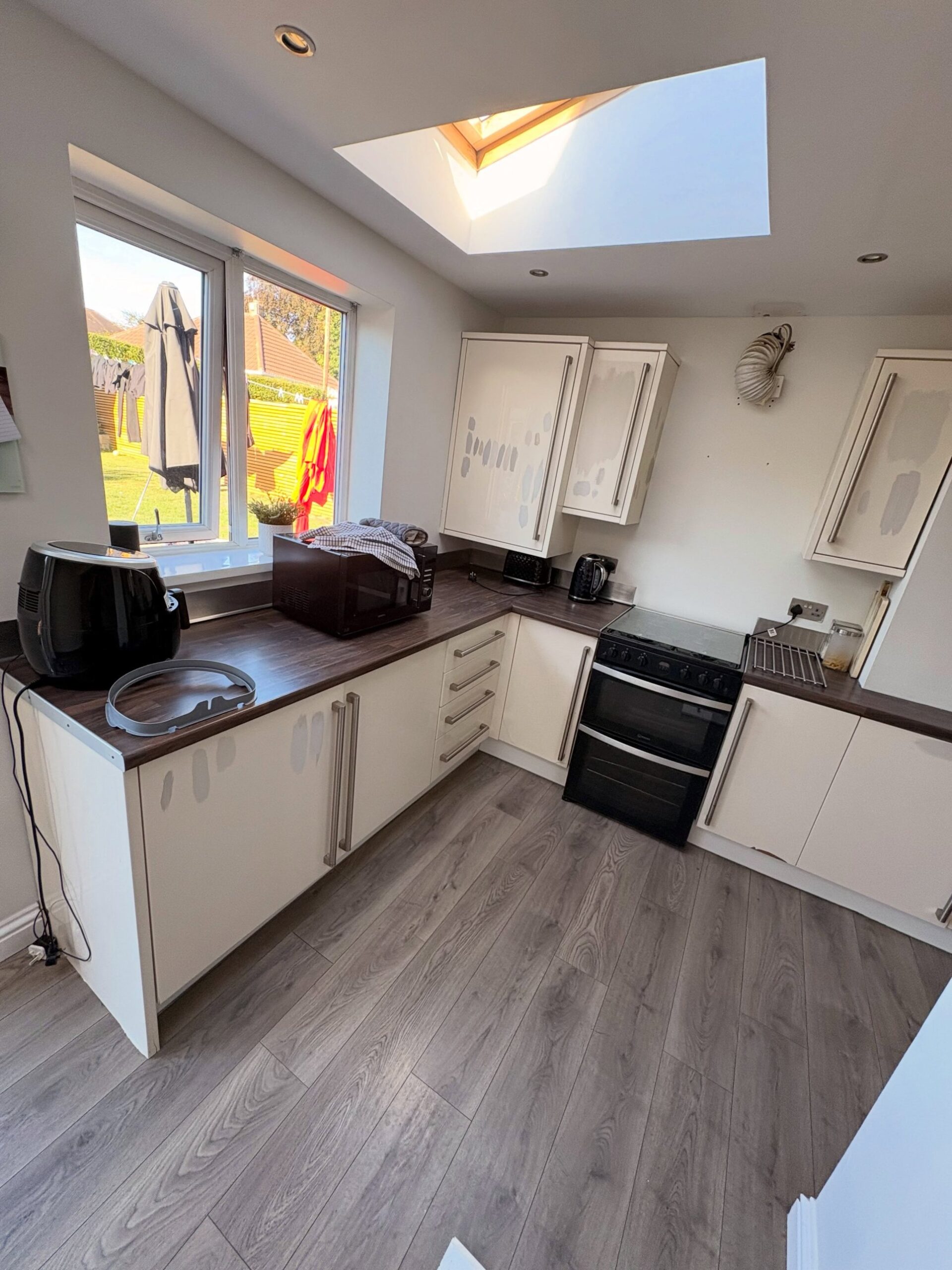U-shaped kitchen with cream cabinets covered in patchy repair marks where vinyl wraps were peeled off, dark walnut counters, long vertical brushed handles, skylight above, laundry drying outside the window.