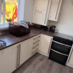 U-shaped kitchen with cream cabinets covered in patchy grey repair marks where failing vinyl wraps were peeled off the doors, dark walnut counters, black oven in the corner, air fryer and microwave on the counter under the back window.