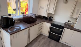 U-shaped kitchen with cream cabinets covered in patchy grey repair marks where failing vinyl wraps were peeled off the doors, dark walnut counters, black oven in the corner, air fryer and microwave on the counter under the back window.