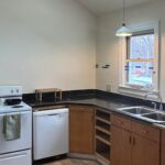 Dated small sloped-ceiling kitchen with a white electric range, white dishwasher, basic brown builder-grade lower cabinets with open bays, dark speckled tile counters, a double stainless sink under the window, a white pendant lamp, and a skylight.