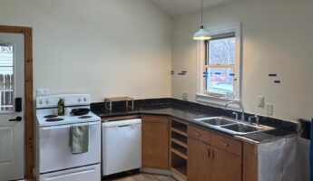 Dated small sloped-ceiling kitchen with a white electric range, white dishwasher, basic brown builder-grade lower cabinets with open bays, dark speckled tile counters, a double stainless sink under the window, a white pendant lamp, and a skylight.
