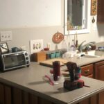 Cramped 1980s kitchen with a dark cherry peninsula jutting into the room, speckled gray laminate counters, dark cherry upper cabinets on the right, a back door on the left, and orange terracotta tile flooring.