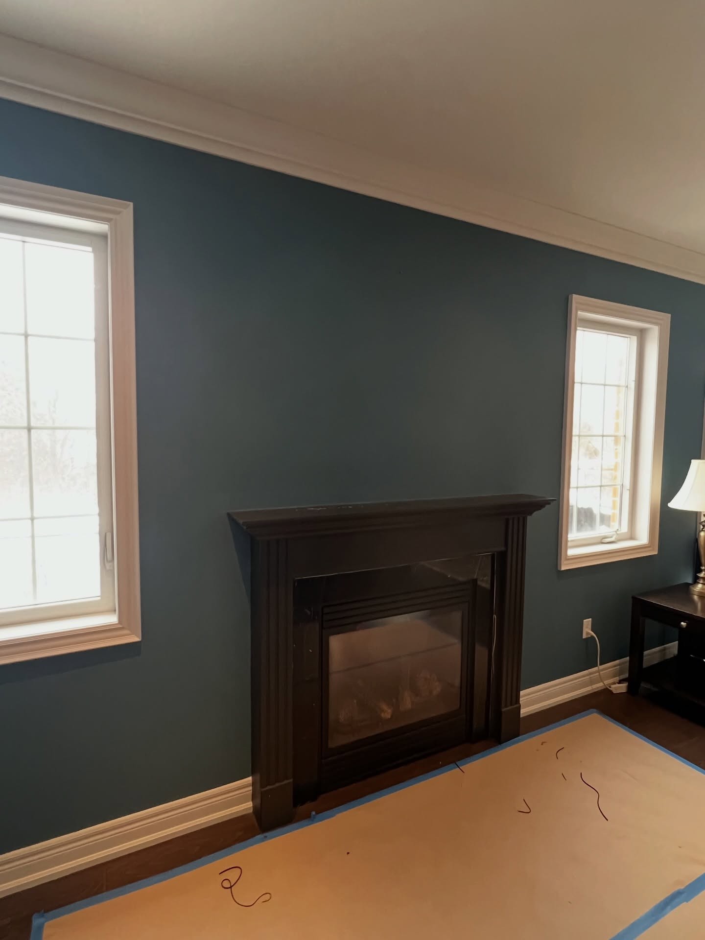Wide room view showing the small dark espresso fireplace on a wall between two windows with a dark side table and lamp to the right, teal walls, white baseboards, and dark hardwood floors.