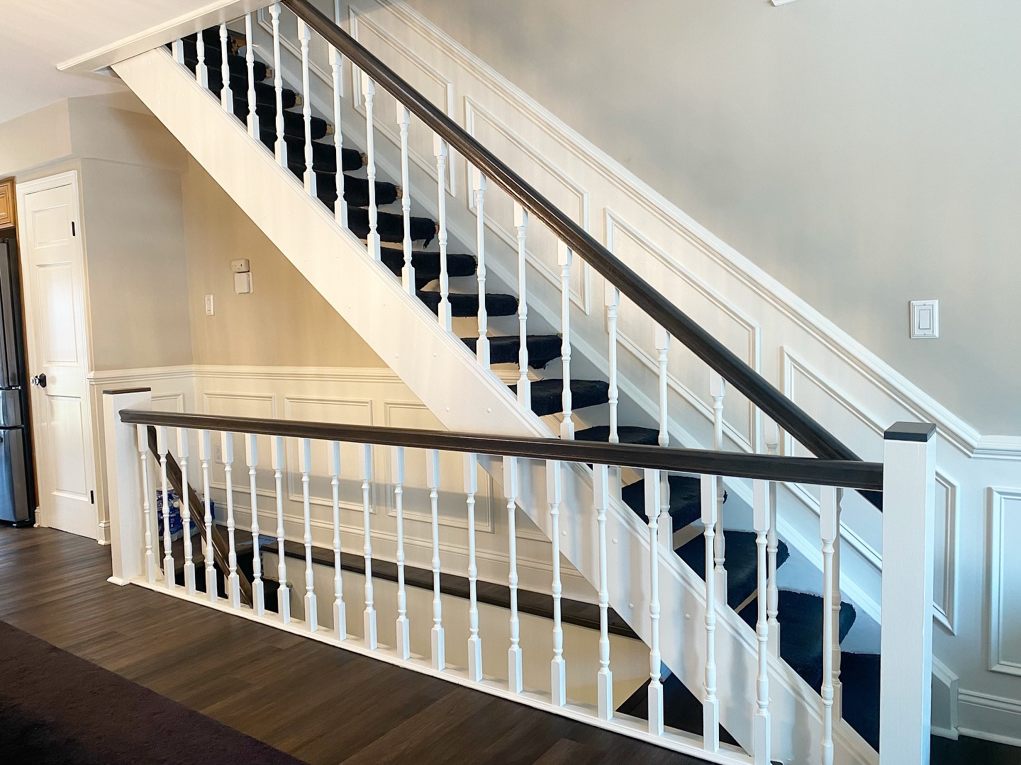 Wide view of a finished staircase with crisp white spindles, custom dark stained handrail, white wainscoting and dark wood floors below.