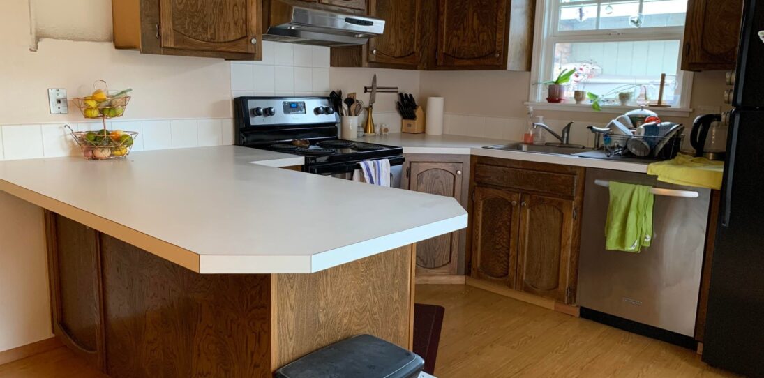 Wide view of a dated 1970s U-shaped kitchen with medium-stained oak cathedral arch cabinets, a white laminate peninsula counter, a black fridge, and a window over the sink.