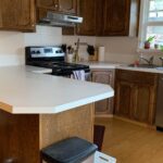 Wide view of a dated 1970s U-shaped kitchen with medium-stained oak cathedral arch cabinets, a white laminate peninsula counter, a black fridge, and a window over the sink.