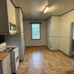 Dated ’90s-style kitchen with teal trim, gray-white cabinets, a small white gas stove, a fluorescent tube light, a steel sink, and beige patterned vinyl flooring before the renovation.