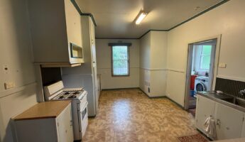 Dated ’90s-style kitchen with teal trim, gray-white cabinets, a small white gas stove, a fluorescent tube light, a steel sink, and beige patterned vinyl flooring before the renovation.
