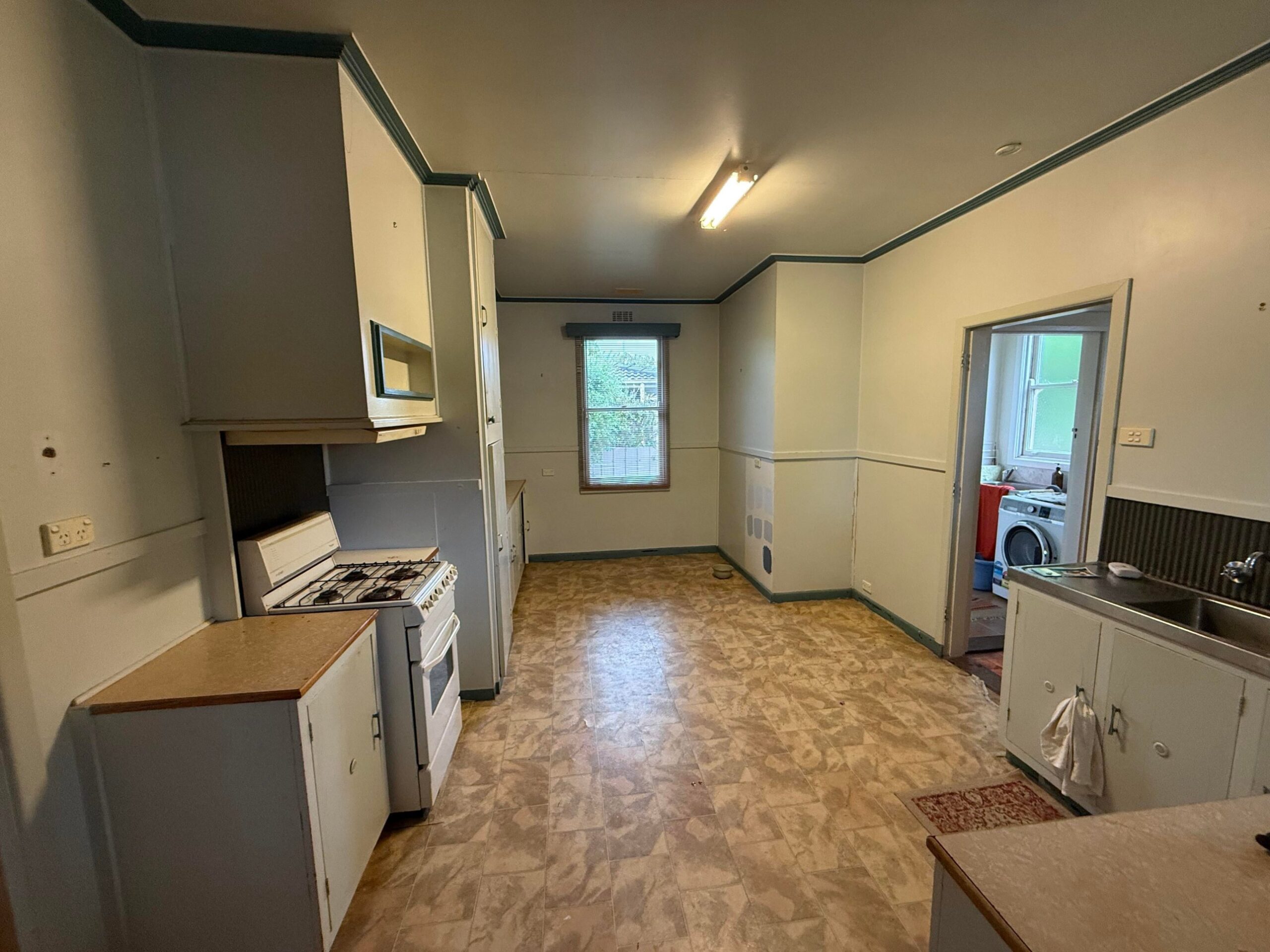 Wide view of an old kitchen with gray-white cabinets, teal trim, a white gas stove, a fluorescent tube light, and beige patterned vinyl flooring.