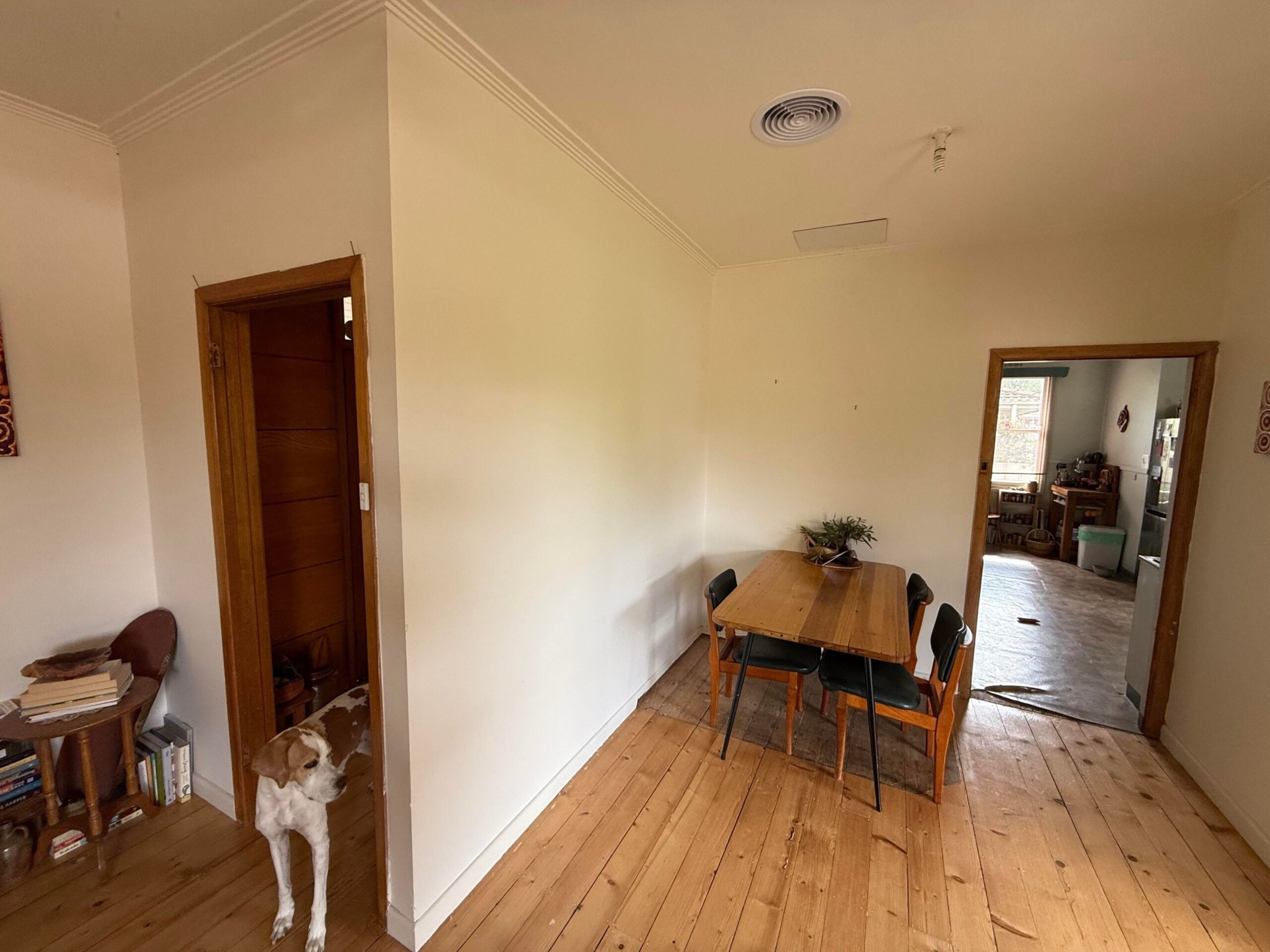 Dining room with pine floorboards, a small wood table with black chairs, a plain white wall, and a single doorway leading into the old kitchen.