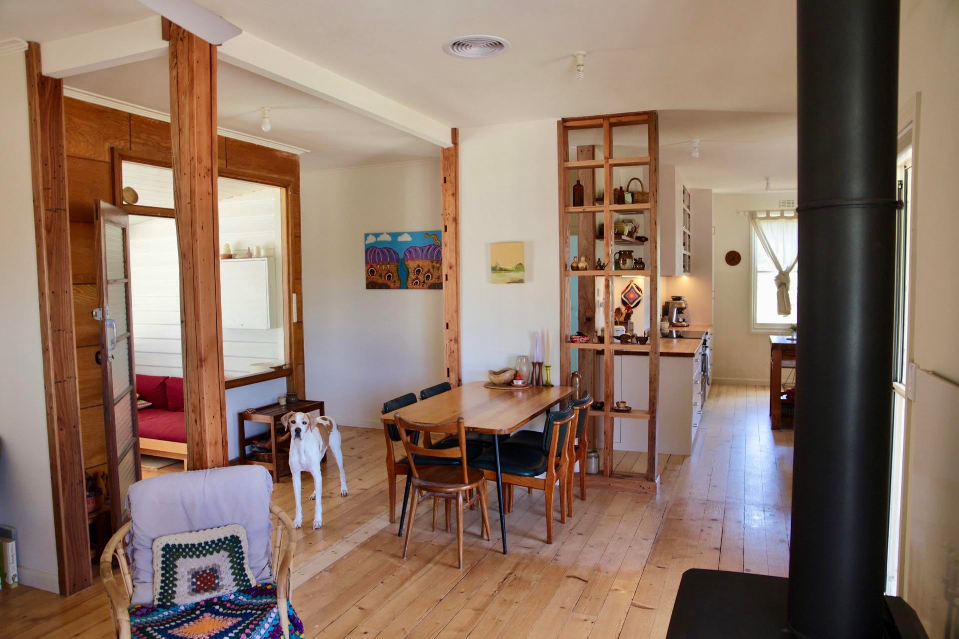 Bright dining room with a wood table, pine floors, and exposed timber posts, plus a stud wall turned into open shelves with pottery that looks into the kitchen.