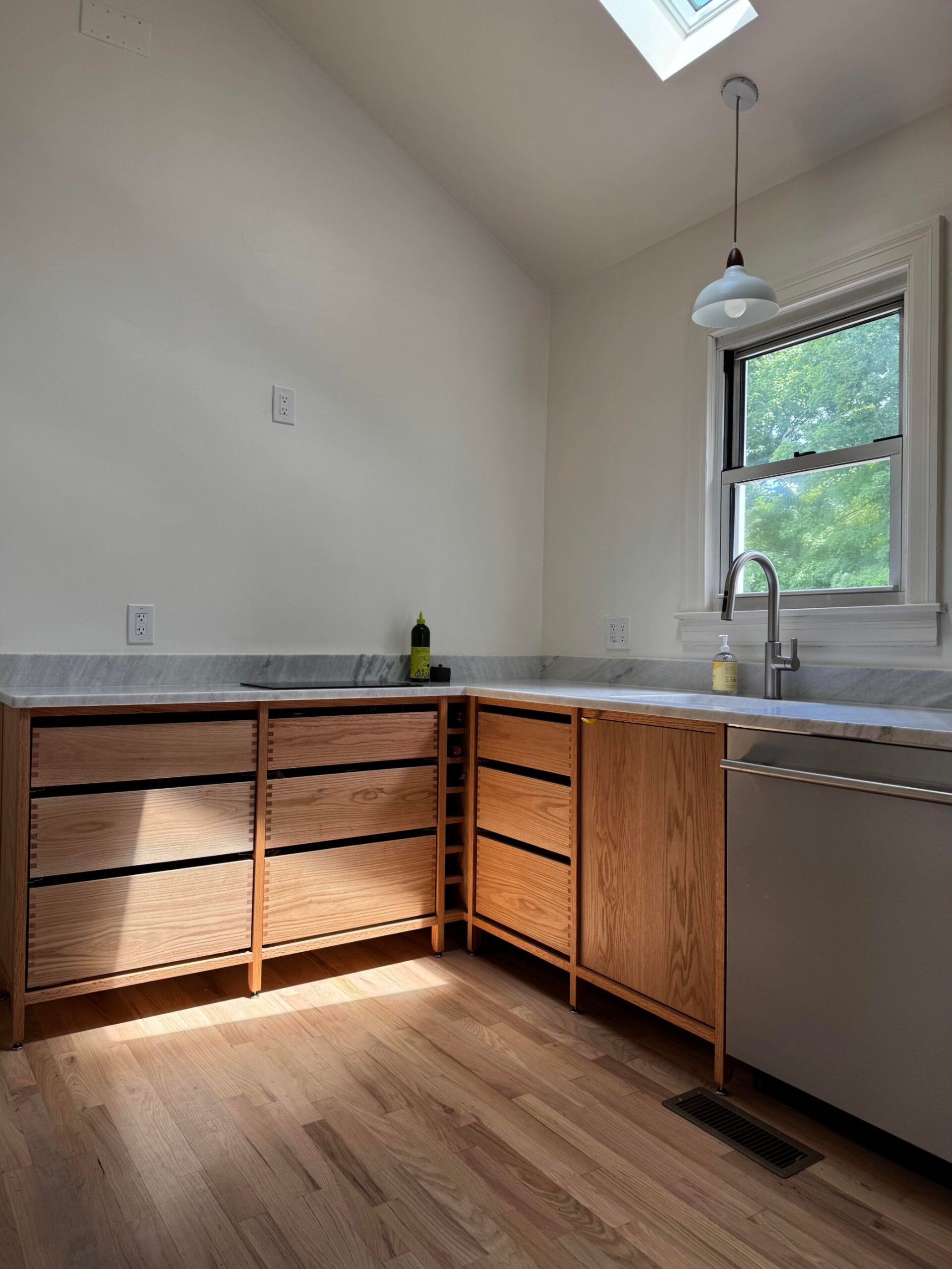 Small L-shaped kitchen with solid red oak lower cabinets on slim steel legs, white marble counter, no upper cabinets, a window above the sink, and a stainless dishwasher.