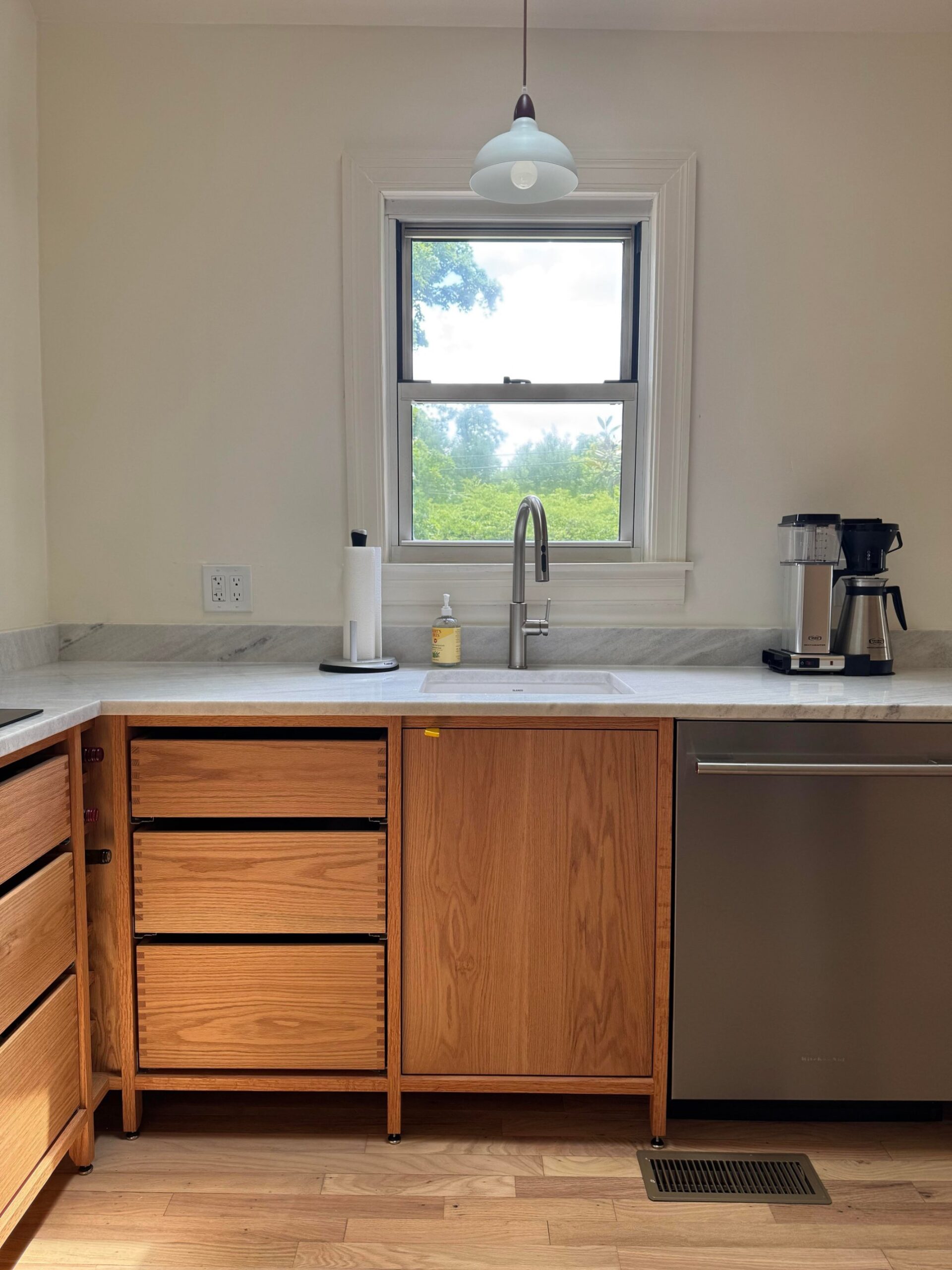 Front view of the sink area with sunlight through the window, white marble counter, stacked oak drawers on the left, a single oak door below the sink, and a stainless dishwasher on the right.