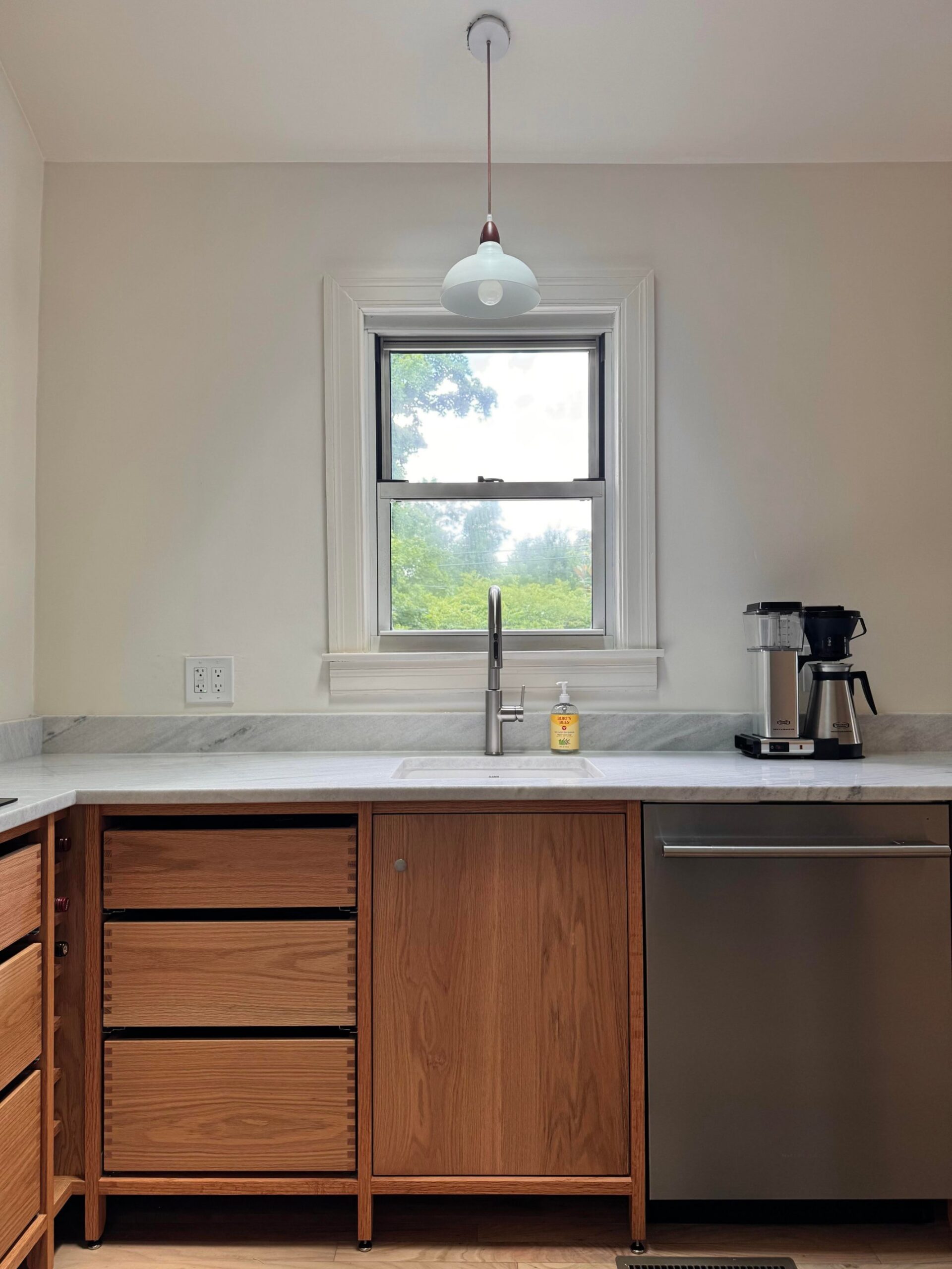 Front view of the finished oak kitchen sink wall with marble counter, undermount sink, stainless faucet, pendant lamp, and a Moccamaster coffee maker on the right.