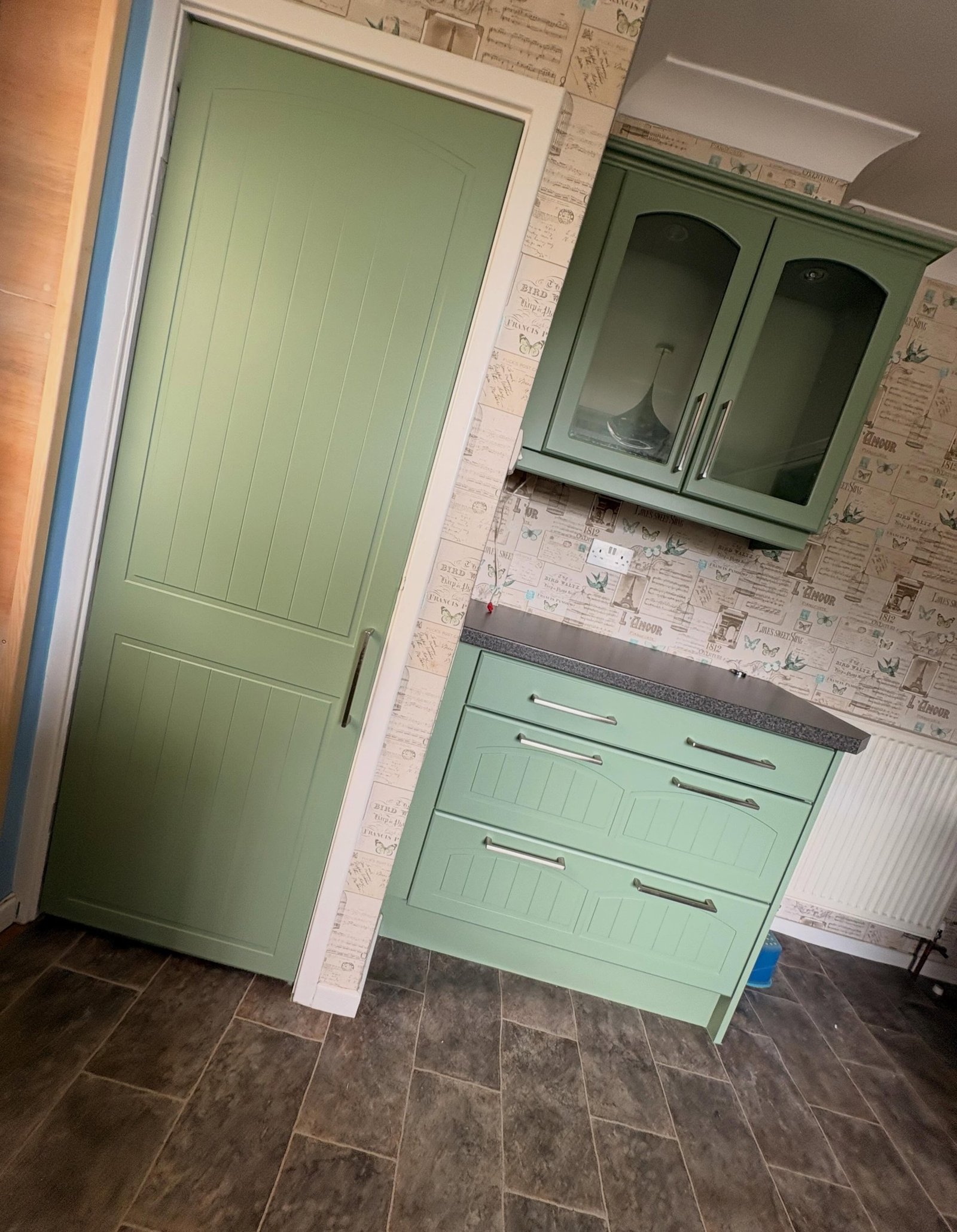 Back view of a UK kitchen during respray prep, with pantry door, wall cabinet, radiator and floor all masked in plastic.