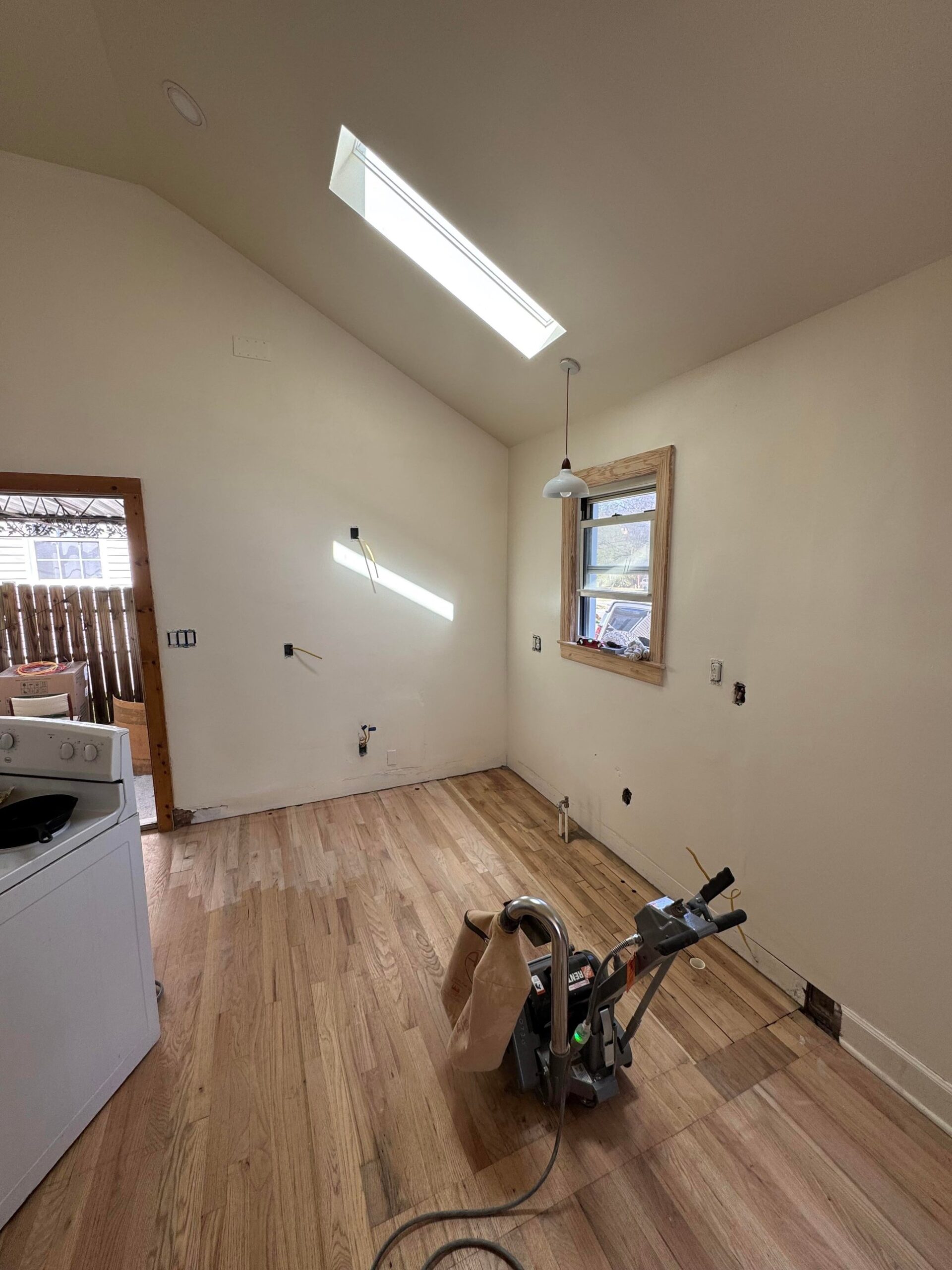 Empty kitchen stripped of cabinets with a drum floor sander in the middle of raw red oak flooring, under a skylight.