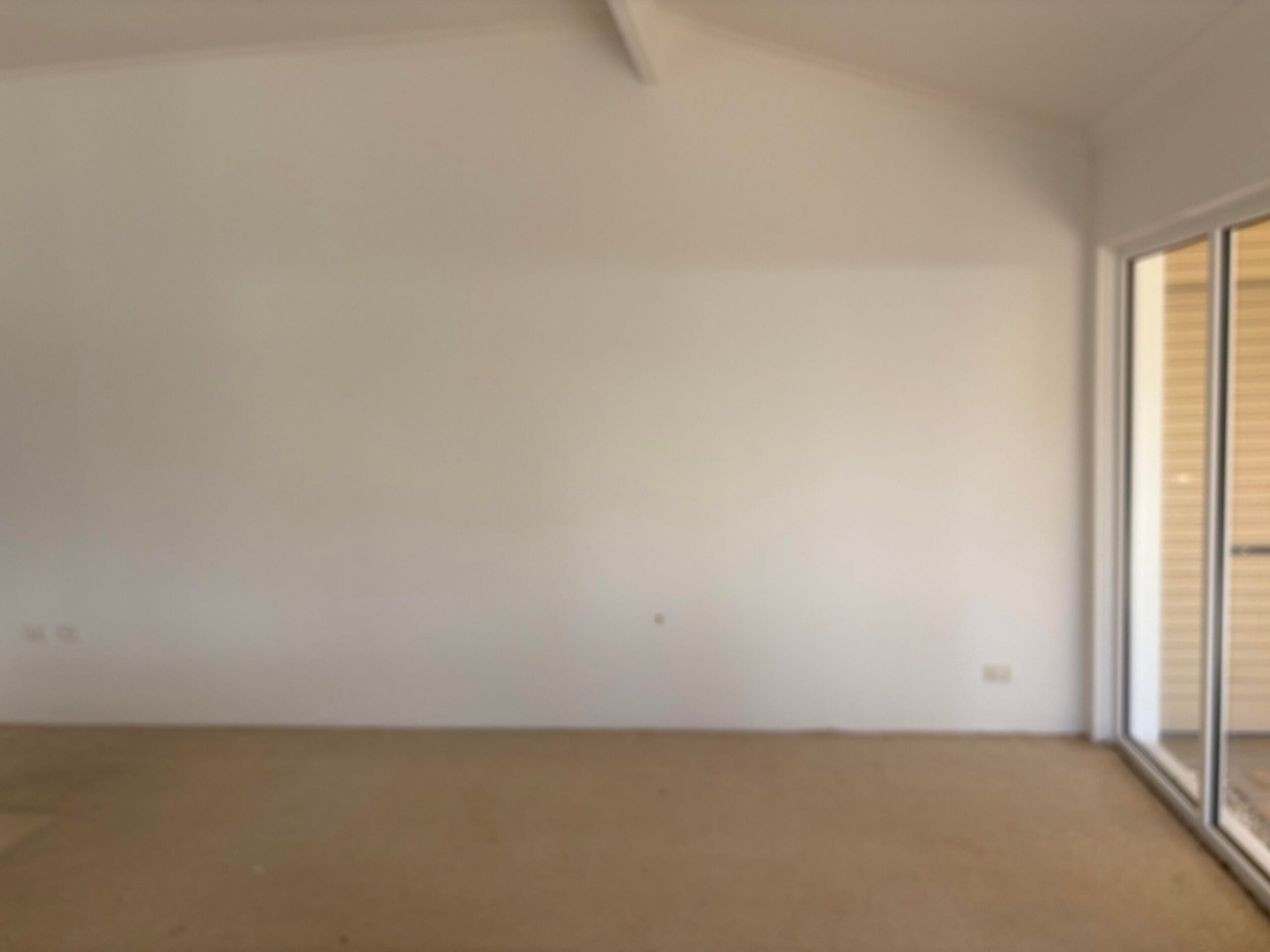 Blank white wall in the original living room with beige carpet, a vaulted ceiling, and a sliding glass door on the right.