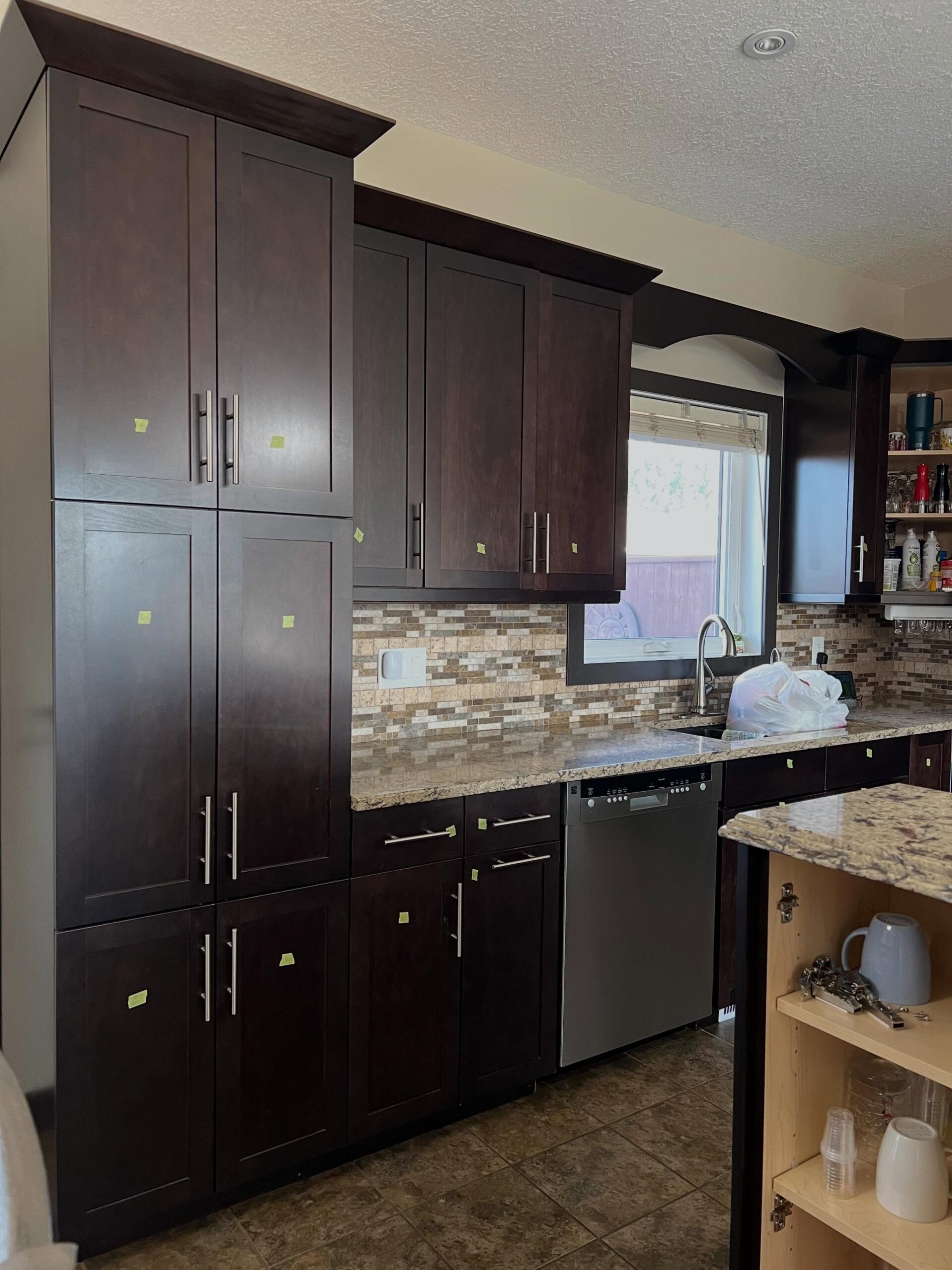 Close view of the dark espresso maple pantry wall before the paint job, with brushed nickel bar pulls, the tumbled stone mosaic backsplash, speckled granite counters, and the stainless dishwasher to the right of the sink.
