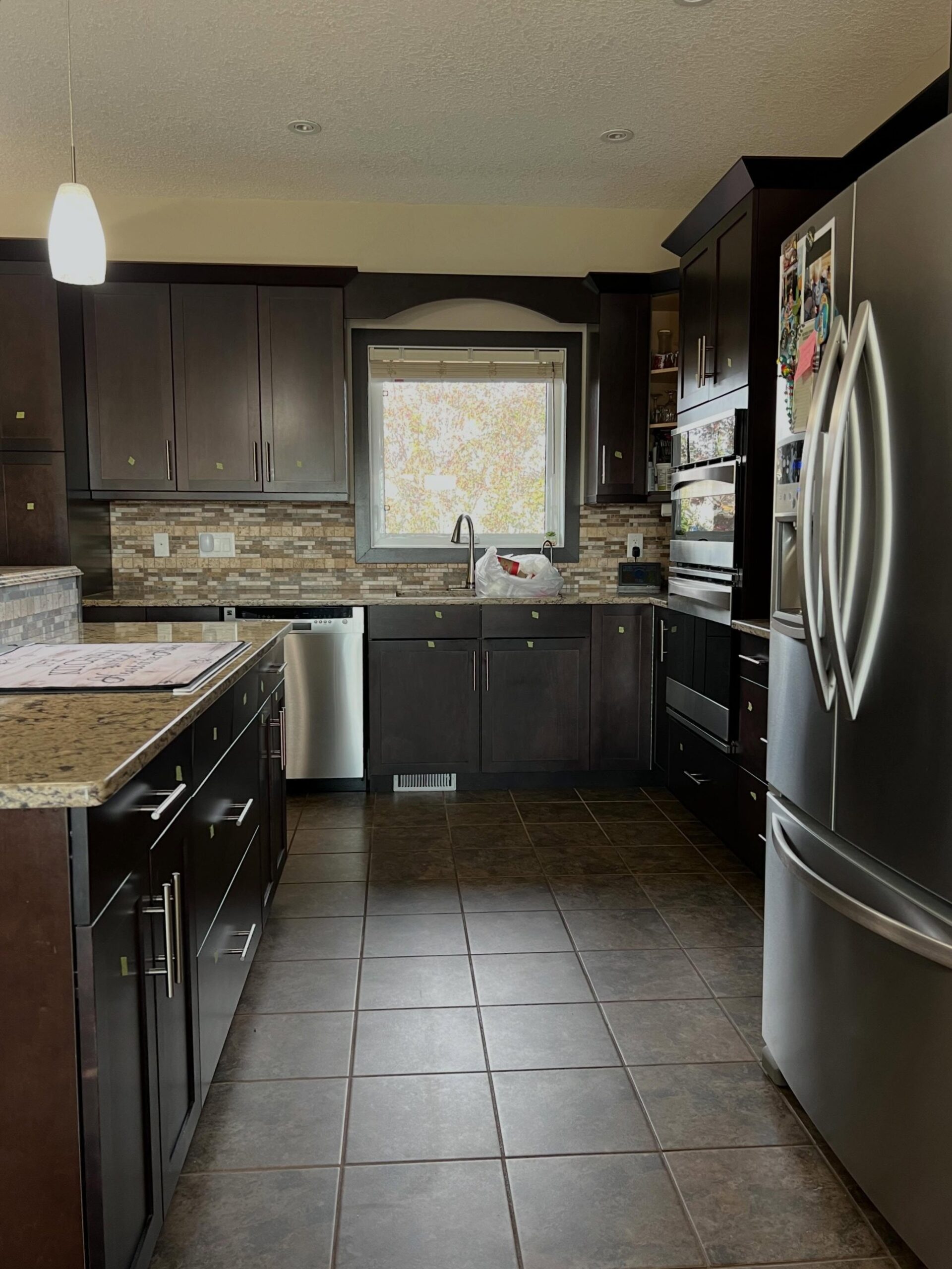 Head-on view of the dark espresso maple kitchen before the paint job, with the arched valance over the sink window, double wall oven to the right, stainless dishwasher to the left, and brown floor tile running toward the camera.