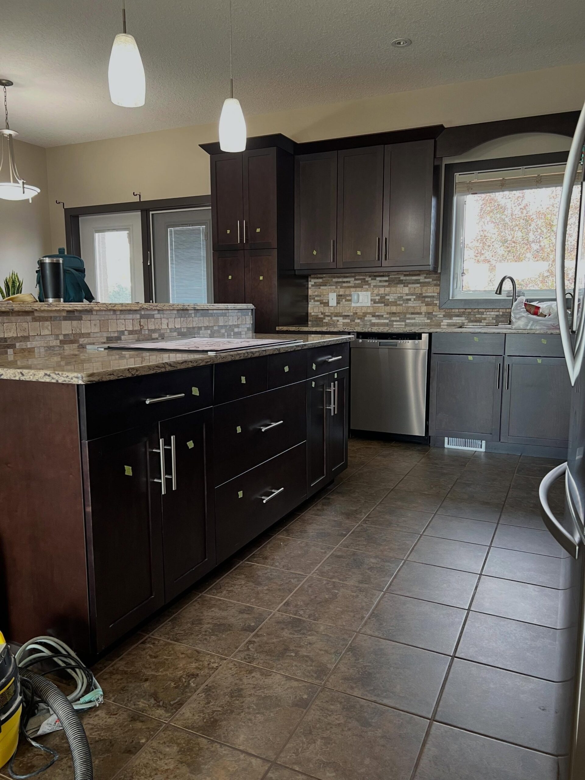 Wide view of the same U-shaped kitchen before the paint job, with dark espresso-stained maple shaker cabinets, brushed nickel bar pulls, gold-and-brown speckled granite counters, a tumbled stone mosaic backsplash, brown ceramic floor tile, and a stainless steel fridge visible on the right.