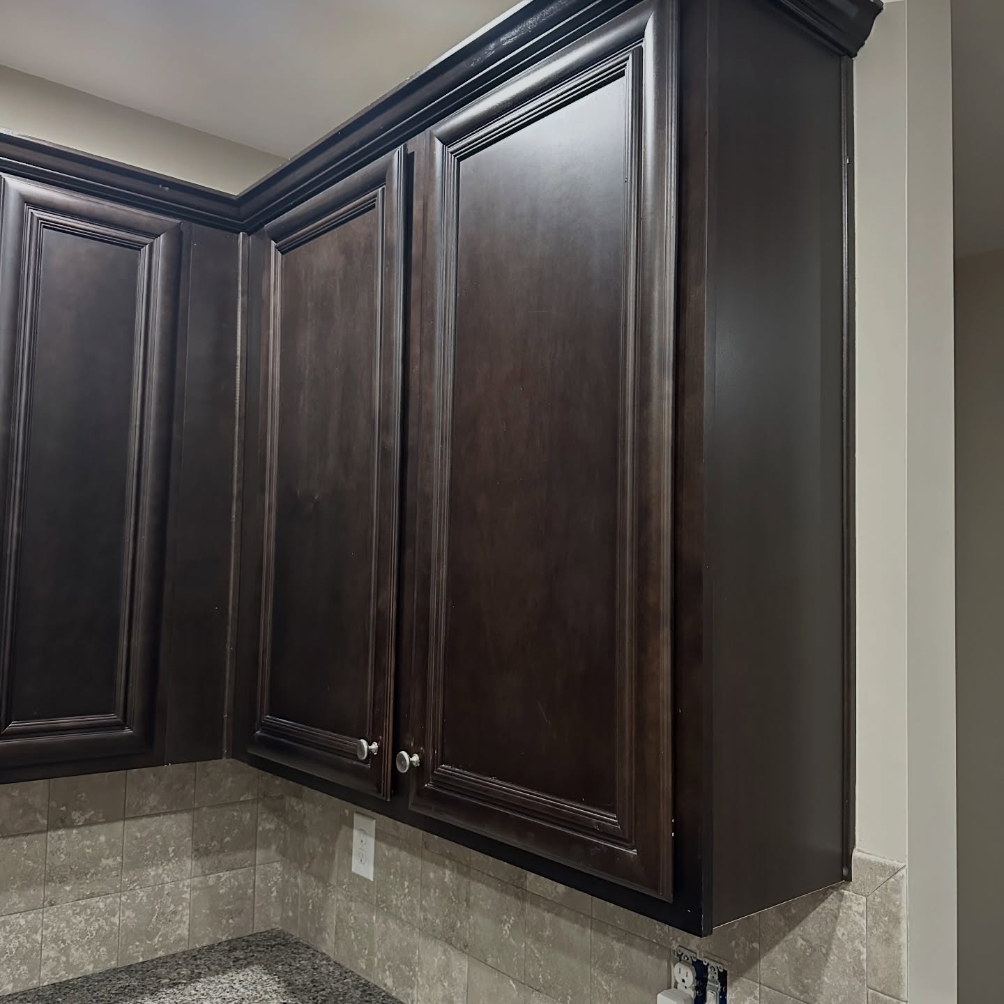 Close-up of the same upper cabinets before paint, in a dark espresso stain, with slightly streaky color and visible shadow lines inside the raised-panel moldings, over the same beige tile backsplash.
