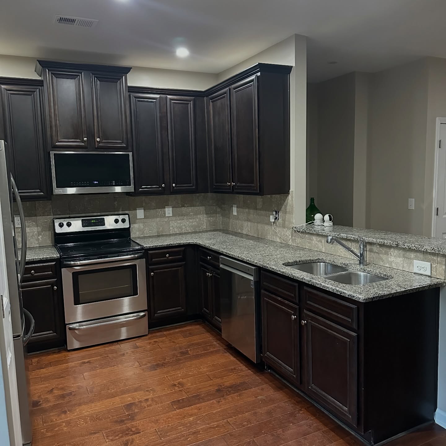 Wide view of the kitchen before paint, with espresso cabinets, speckled gray and white granite counters, a stainless dishwasher and stove, a beige tile backsplash, and a small green bottle on the counter.