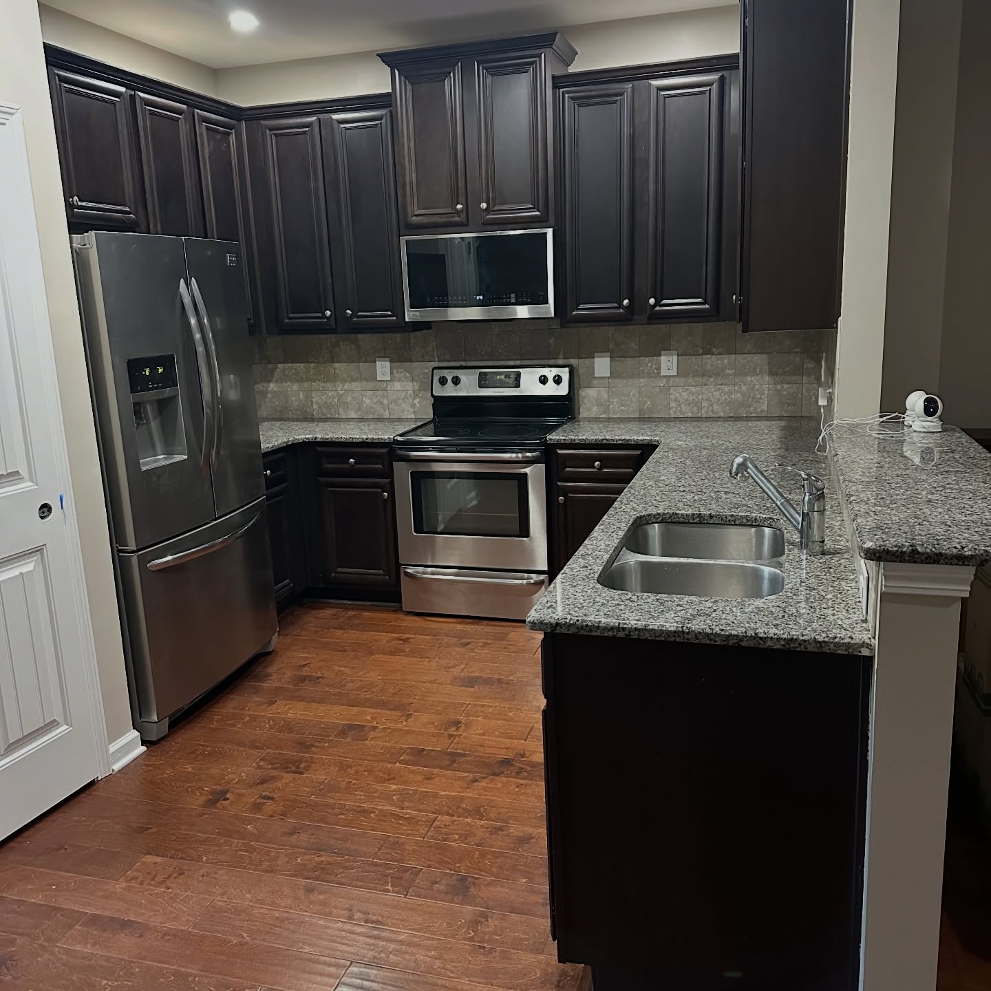 U-shaped kitchen before the paint job, with dark espresso raised-panel cabinets, speckled gray and white granite counters, a beige tile backsplash, stainless steel appliances, and a small security camera sitting on the counter.