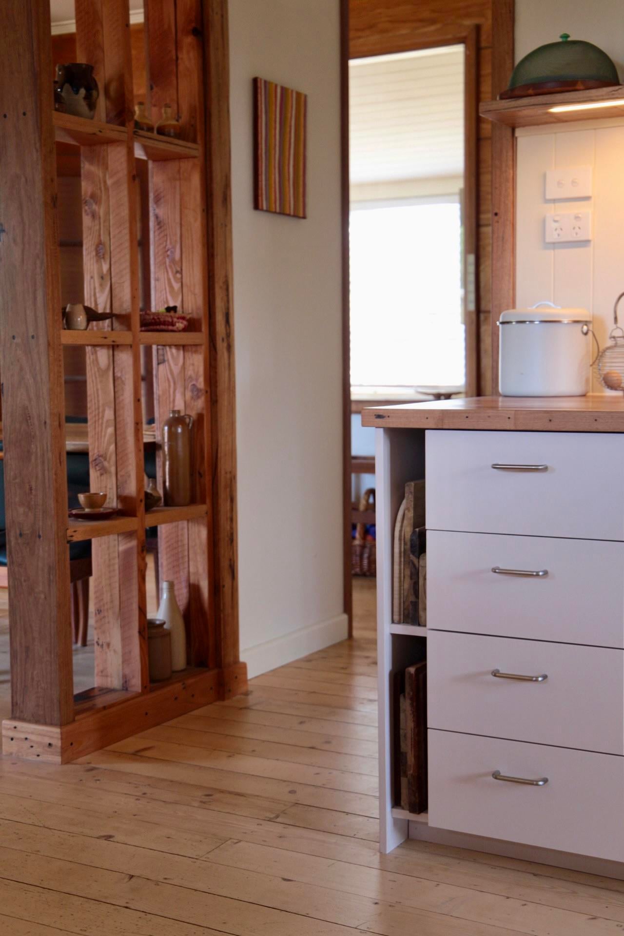 Close view of exposed wood studs with small shelves holding pottery and bottles, next to white kitchen drawers with cutting boards tucked in an end slot.