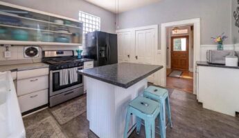 Small 1960s-style kitchen inside a 122-year-old Victorian home, with a black side-by-side refrigerator in front of a glass block window, white laminate counters, teal metal stools, and a tile floor.