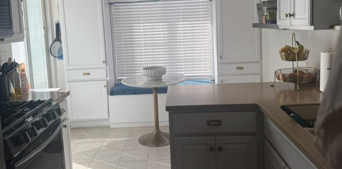 Cramped galley kitchen before renovation, with a short tan laminate peninsula counter jutting into the walkway, a tulip bistro table squeezed in front of a window seat with blinds, worn beige tile flooring, and a black gas stove on the left wall.