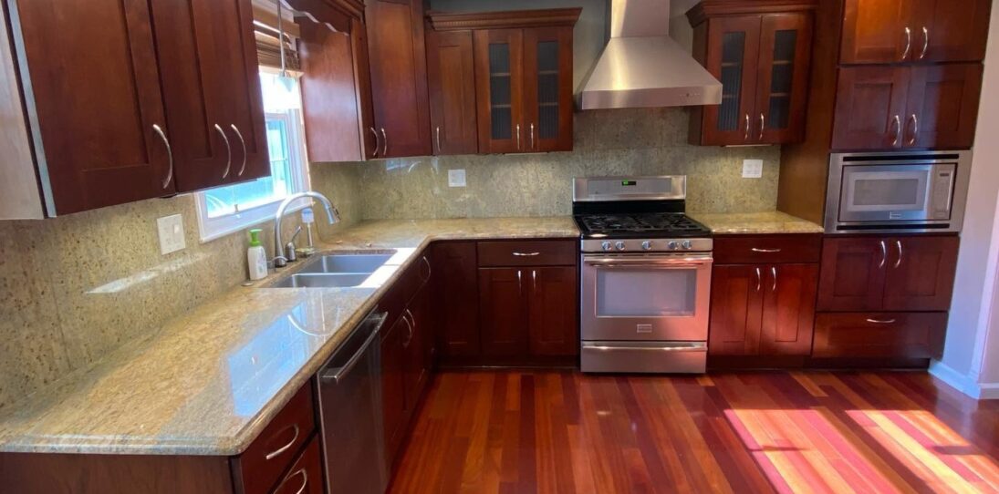 Dark cherry wood kitchen with raised panel cabinets, speckled gold and tan granite counters, stainless steel range and hood, and glossy red Brazilian cherry hardwood floors, shown before being painted Sherwin-Williams Extra White.