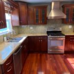 Dark cherry wood kitchen with raised panel cabinets, speckled gold and tan granite counters, stainless steel range and hood, and glossy red Brazilian cherry hardwood floors, shown before being painted Sherwin-Williams Extra White.