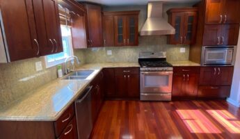 Dark cherry wood kitchen with raised panel cabinets, speckled gold and tan granite counters, stainless steel range and hood, and glossy red Brazilian cherry hardwood floors, shown before being painted Sherwin-Williams Extra White.