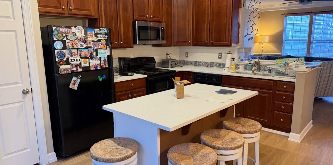 Wide 16:9 view of a kitchen before renovation, with orange cherry wood cabinets, a large white quartz island surrounded by four round rush-seat stools, a black fridge covered in travel magnets, a stainless microwave, a black stove, and light oak LVP flooring running throughout.