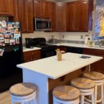 Wide 16:9 view of a kitchen before renovation, with orange cherry wood cabinets, a large white quartz island surrounded by four round rush-seat stools, a black fridge covered in travel magnets, a stainless microwave, a black stove, and light oak LVP flooring running throughout.