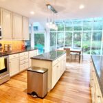 Wide view of a dated 1990s kitchen with peeling cream shabby chic cabinets, orange botanical tile backsplash, brown granite countertops, a stainless microwave hood, a small painted island, and large arched sunroom windows at the end of the room.