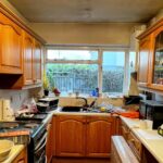 Cramped U-shaped galley kitchen with worn orange oak cabinets, white laminate counters cluttered with caulk tubes and a Nikotek box, a double window above the sink looking onto a wooden garden fence, a textured ceiling, and glass-front upper cabinets on the right wall.