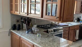 Wide view of a kitchen before renovation with honey oak cabinets, mullion glass upper doors with pinecone pulls, speckled granite counters, a stainless gas range, and an arched wine lattice cabinet on the right.